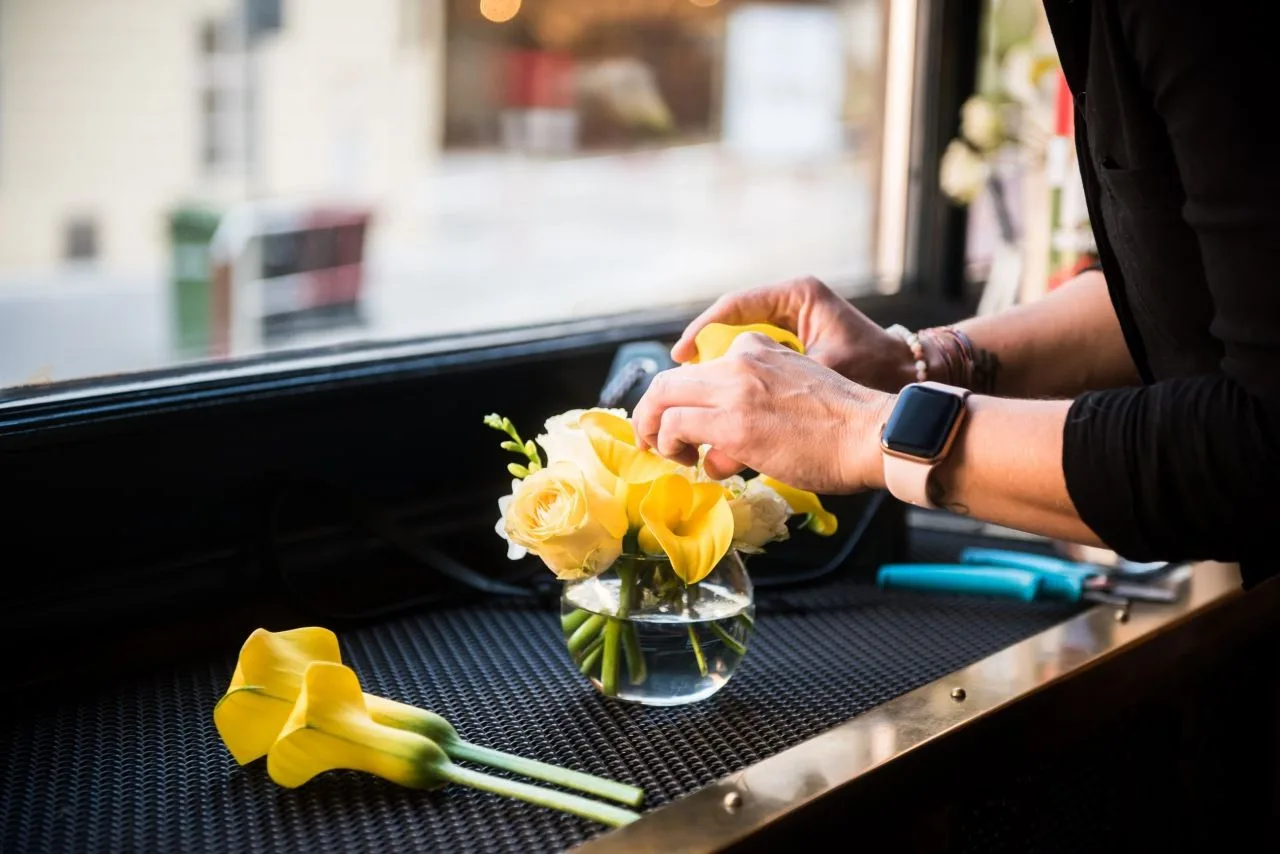 Ruce floristky aranžují žluté kaly a růže do kulaté skleněné vázy u okna